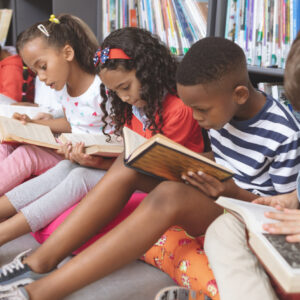 Side view of mixte ethnicity school kids sitting on cushions and studying over books in a library at school against bookshelfs in background